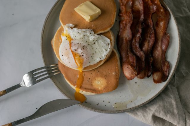 Top view of delicious pancakes with poached egg with seasoning and butter piece near sliced fried bacon on plate near fork and knife near napkin on white table
