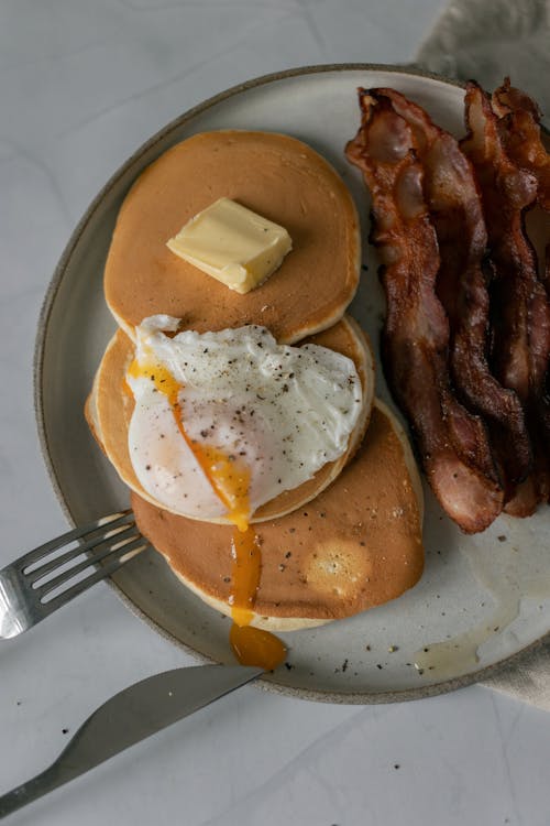 Free From above of pancakes with poached egg with seasoning and butter piece near sliced fried bacon on plate near fork and knife on white table Stock Photo