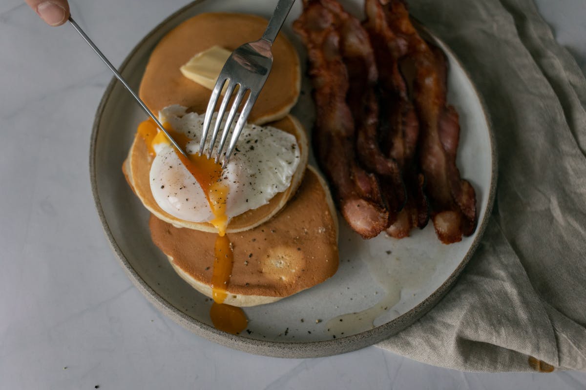 From above of unrecognizable person cutting poached egg served on pancakes with roasted bacon during breakfast in kitchen