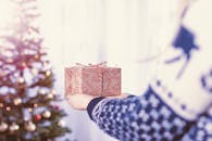 在舒适的室内，一个穿着毛衣的人正拿着包装好的礼物站在装饰好的圣诞树旁