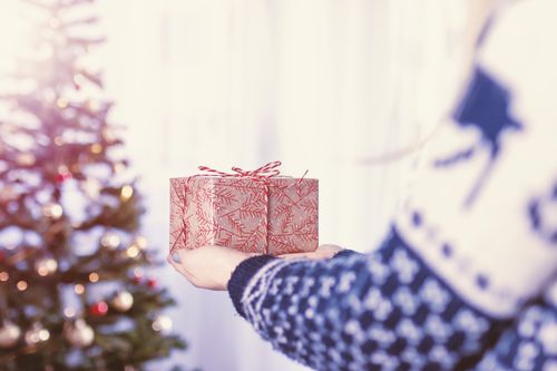 Free A person in a sweater holds a wrapped gift near a decorated Christmas tree in a cozy indoor setting. Stock Photo