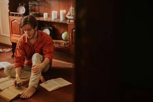Free A young man reading books by candlelight in a vintage room, creating a warm, introspective atmosphere. Stock Photo