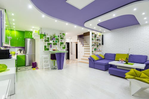 Interior of modern spacious illuminated house with comfortable purple sofa in living room and bright green furniture in kitchen area