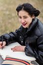 Positive Asian female student with makeup in warm jacket taking notes in copybook while sitting on grassy ground on blurred background