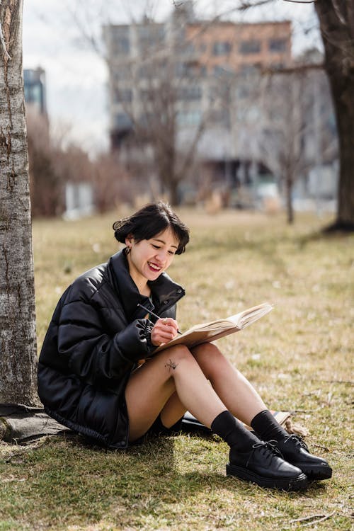 Full body of smiling Asian female student taking notes in workbook while sitting on grassy lawn against residential buildings in town
