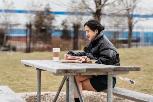 Side view of cheerful Asian female student in jacket writing in copybook while sitting at wooden table with takeaway coffee against blurred background
