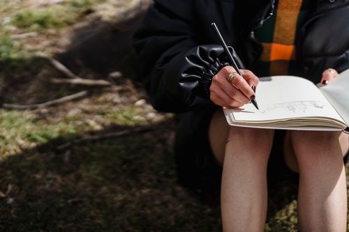 Free Crop unrecognizable female in black jacket drawing with pencil in workbook while sitting on meadow Stock Photo