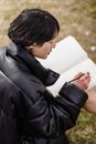 From above side view of young Asian female in black jacket drawing in workbook on lawn