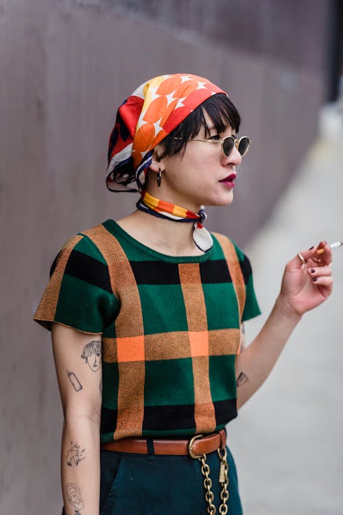 Free Stylish young ethnic female in checkered t shirt and sunglasses with cigarette looking away on urban pavement Stock Photo