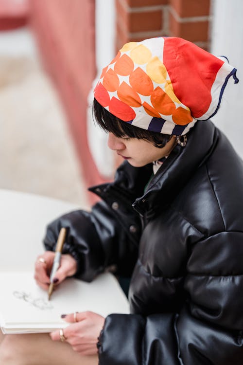 From above side view of young ethnic female in bandana drawing in workbook on street