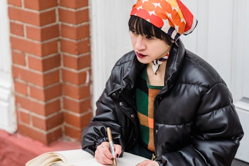 Pensive young ethnic female in padded jacket with pen and workbook preparing for exam while looking away in town