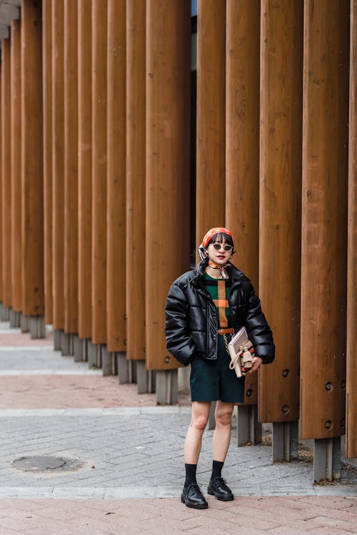 Confident young Asian female in padded jacket and sunglasses standing with hand in pocket on city walkway against colonnade
