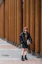 Confident young Asian female in padded jacket and sunglasses standing with hand in pocket on city walkway against colonnade
