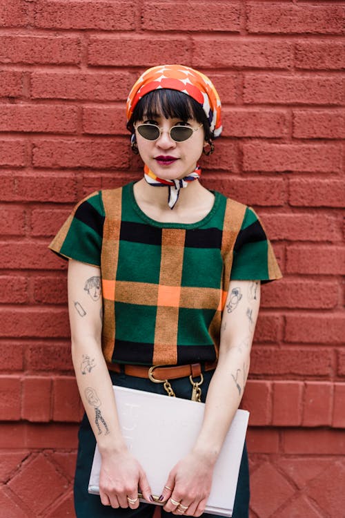 Cool young ethnic woman in checkered t shirt and sunglasses with textbook looking at camera against brick wall