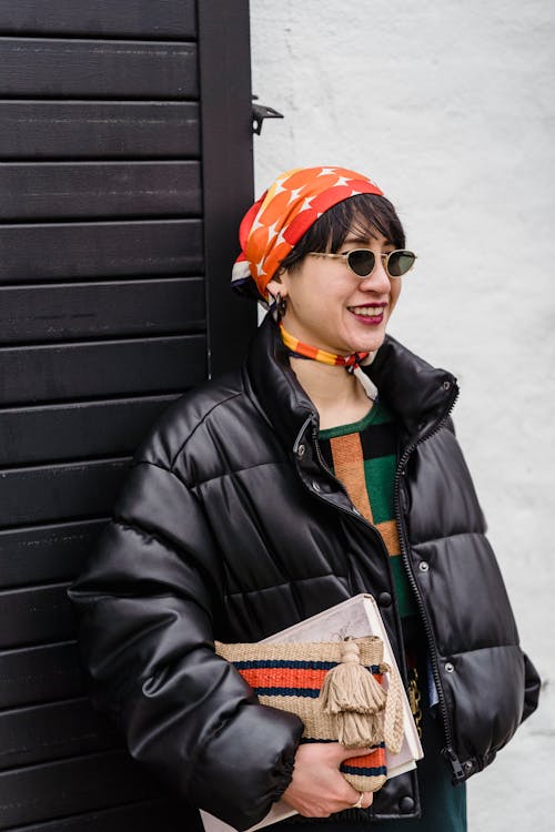 Free Smiling cool ethnic woman in padded jacket and bandana standing with book and handbag against wall Stock Photo
