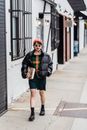 Cool young ethnic woman with book and hand in pocket strolling on walkway in city