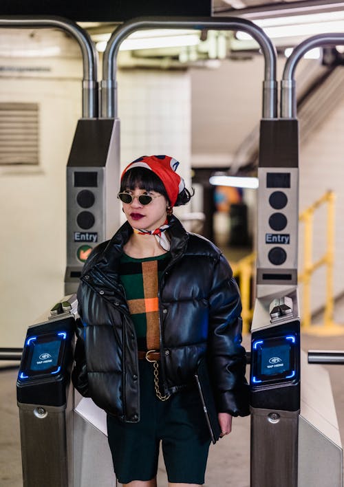 Free Trendy young ethnic female in bandana and padded jacket with hand in pocket looking away while passing through turnstile Stock Photo