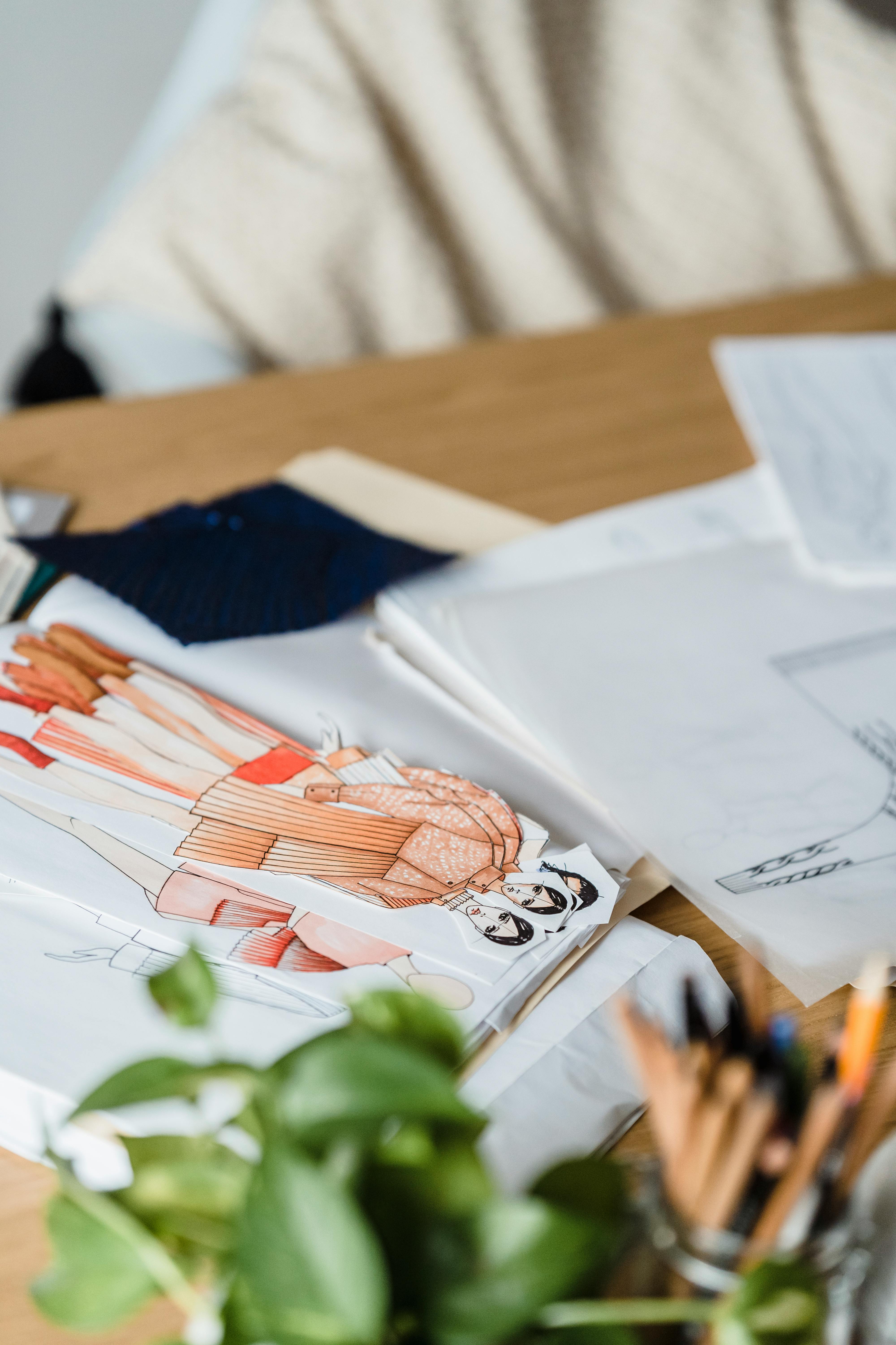 Colorful creative cutout drafts of modern outfits placed on wooden table with papers and green plants in modern light workshop