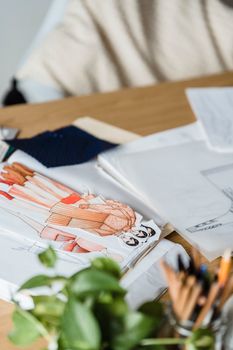 Colorful creative cutout drafts of modern outfits placed on wooden table with papers and green plants in modern light workshop