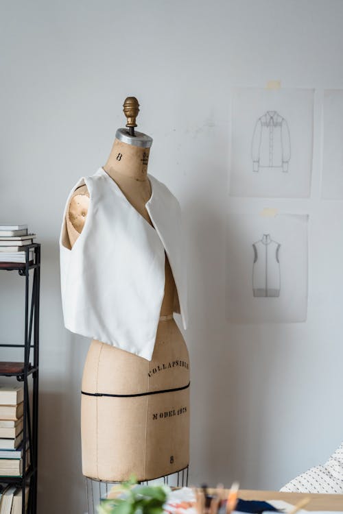 Mannequin in white vest at white wall with drafts of clothes in light room of modern atelier with books