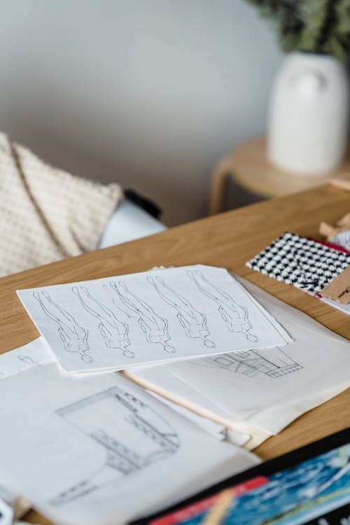 Papers with creative drafts placed on wooden table with sewing patterns and samples of textile in light room of atelier