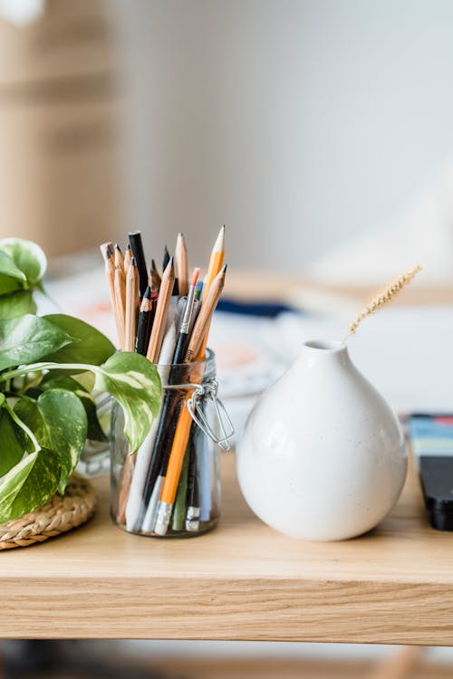 Heap of pencils in glass jar placed near ceramic vase with twig and green plant in light workshop on blurred background