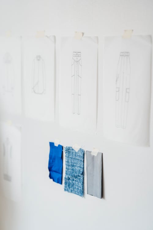 Papers with sketches of stylish garments attached on white wall with tape near samples of colorful fabric in light modern atelier