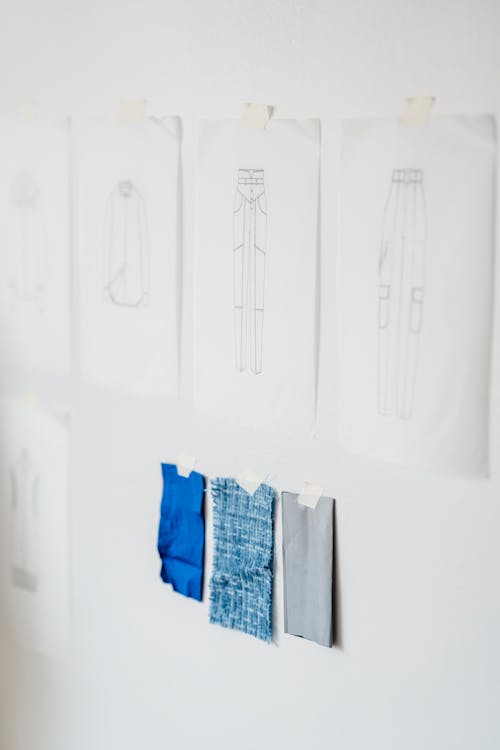 Free Papers with sketches of stylish garments attached on white wall with tape near samples of colorful fabric in light modern atelier Stock Photo