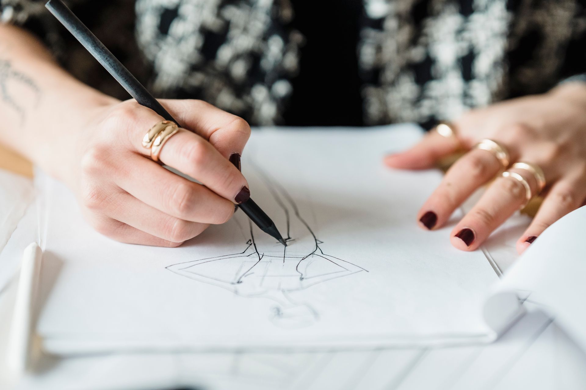 Crop unrecognizable female with rings drawing draft of modern garment while sitting at table with papers in light modern workshop