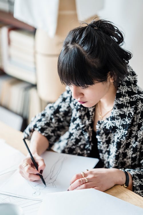 Concentrated female designer drawing draft on paper while sitting at table in modern light workplace with books on blurred background