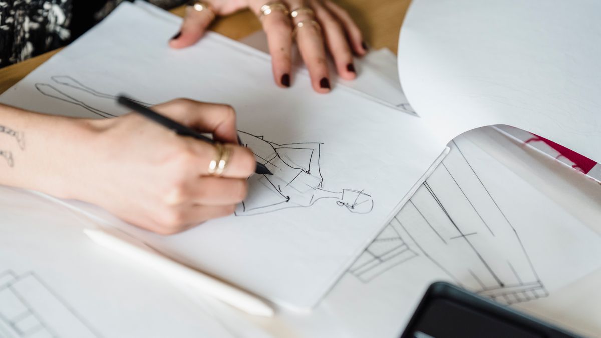 From above of crop anonymous female designer drawing draft of stylish garment at table with papers in modern light workshop