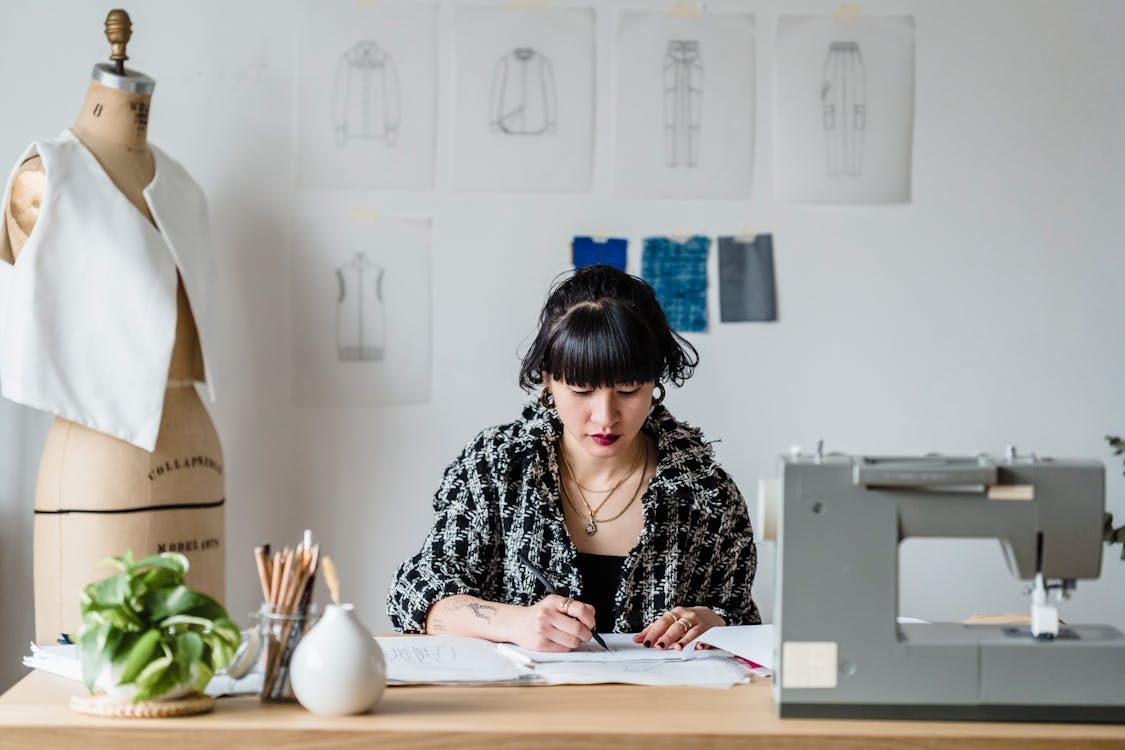 Concentrated ethnic woman drawing sketch while sitting at table near sewing machine and dummy