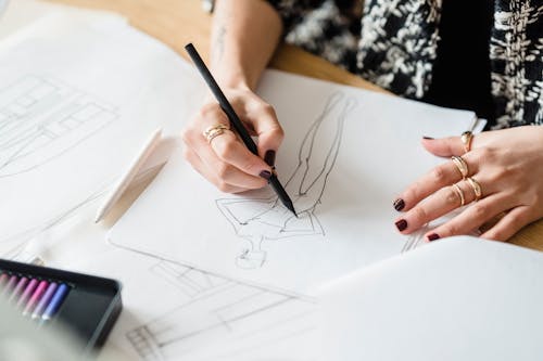 Crop unrecognizable female designer in golden rings drawing human figure with pencil on paper sheet at desk