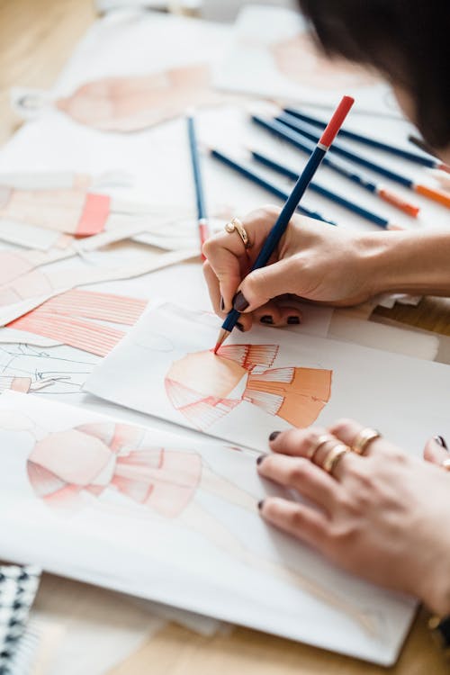 Crop anonymous female illustrator with pencil coloring sketch of wear on human figure on paper sheet at desk