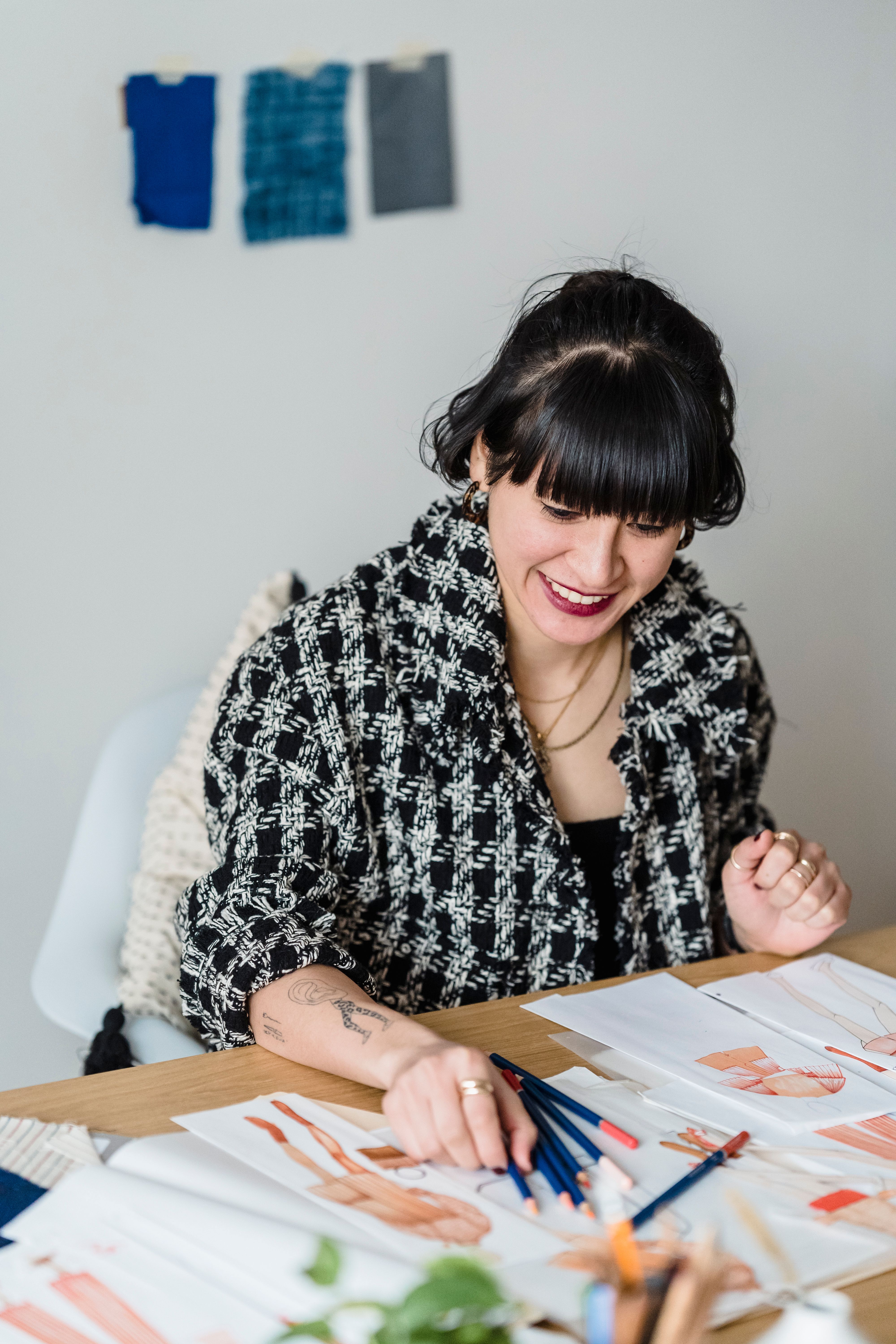 Cheerful young tattooed ethnic female illustrator sitting at table with pencils and clothing sketches while working on project