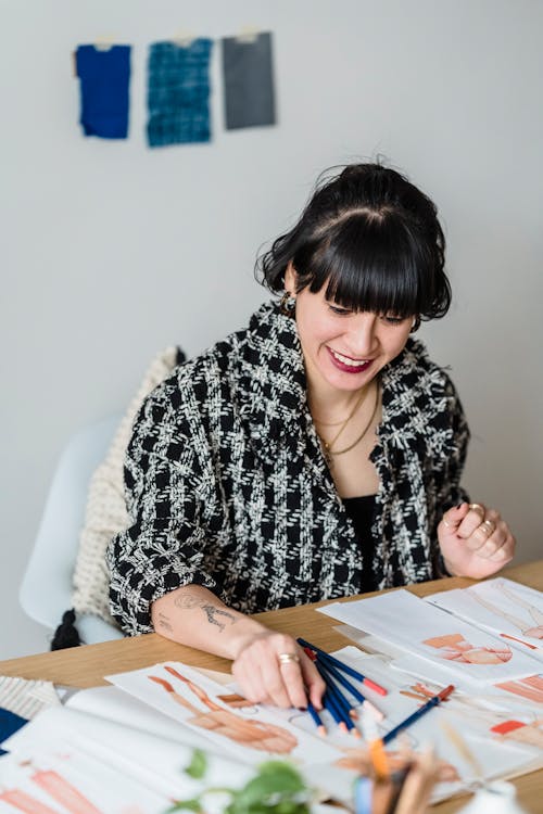 Smiling Asian designer with illustrations and colored pencils at desk