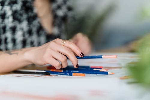 Crop anonymous tattooed female illustrator with collection of colored pencils and paper illustrations at home desk