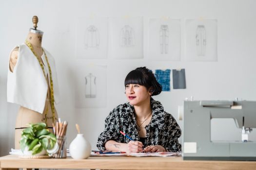 Smiling Asian tailor with illustration against sewing machine in workroom