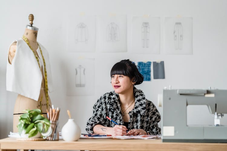 Smiling Asian Tailor With Illustration Against Sewing Machine In Workroom