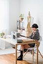 Pensive young ethnic female tailor touching chin while sitting with crossed legs at desk with sewing machine and sketches in atelier