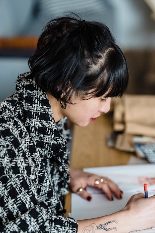 Free Side view of focused young ethnic female illustrator with pencil and tattoo coloring sketch while working at desk Stock Photo
