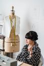Dreamy young Asian female tailor in checkered clothes sitting at table while looking away against dummy in workroom