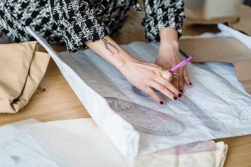 Crop unrecognizable tattooed female tailor measuring paper with curved ruler against fabric on table in atelier