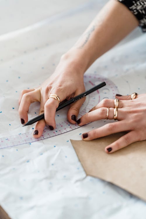 From above of crop anonymous seamstress with pencil measuring paper and cardboard template with curved ruler in workroom