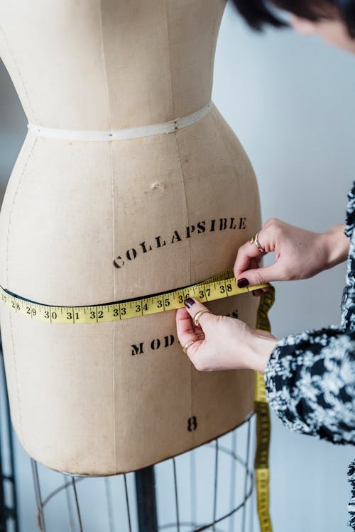Crop unrecognizable female dressmaker with flexible ruler measuring hip line of dummy while working in studio