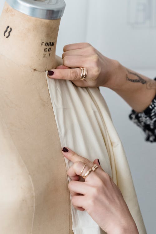 Crop anonymous tattooed female designer in rings measuring cloth on dummy while working in studio