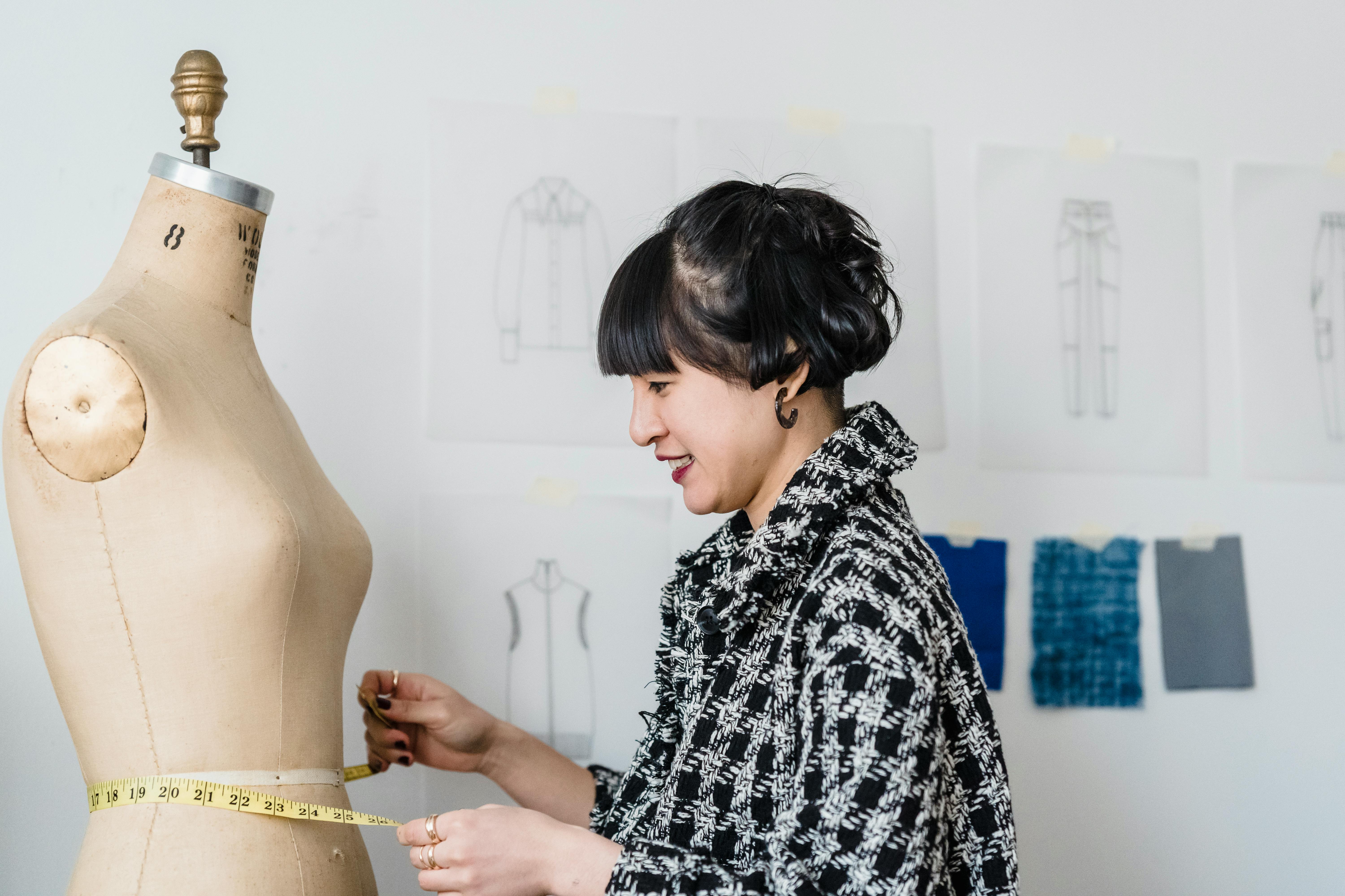 smiling asian tailor with tape measuring mannequin in workshop