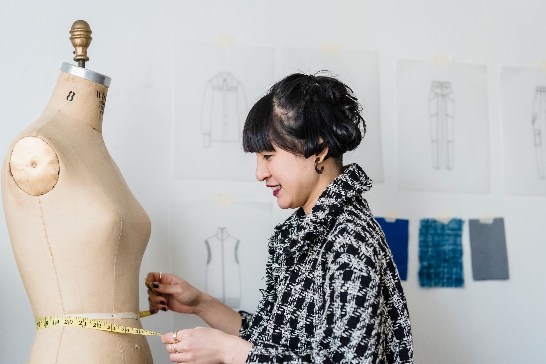 Side view of content young ethnic female dressmaker with flexible ruler measuring waist of dummy in workroom
