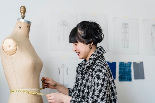 Side view of content young ethnic female dressmaker with flexible ruler measuring waist of dummy in workroom