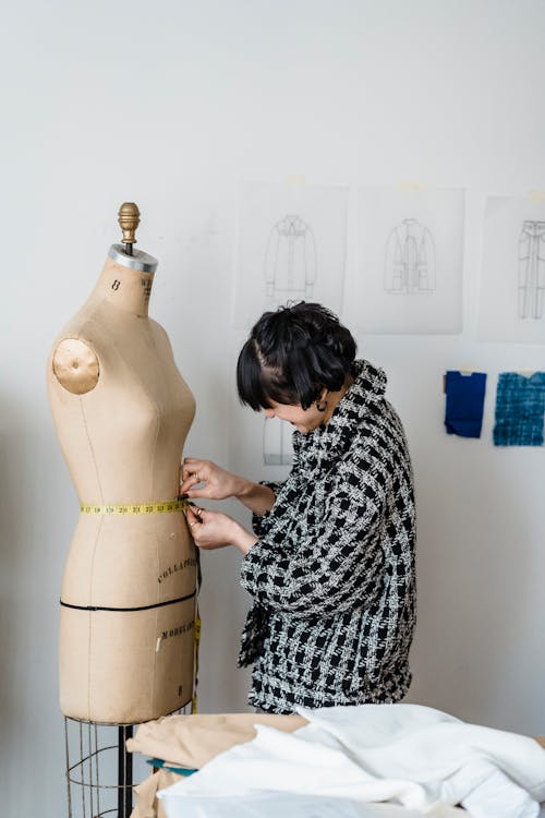 Side view of female tailor in checkered outfit measuring waist of dummy with flexible ruler in atelier